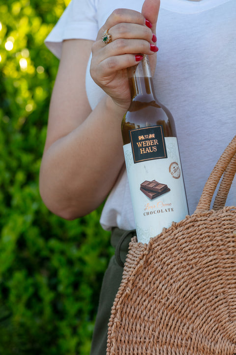 Mulher segurando garrafa de Licor Creme Weber Haus de Chocolate com uma bolsa de palha em ambiente externo.